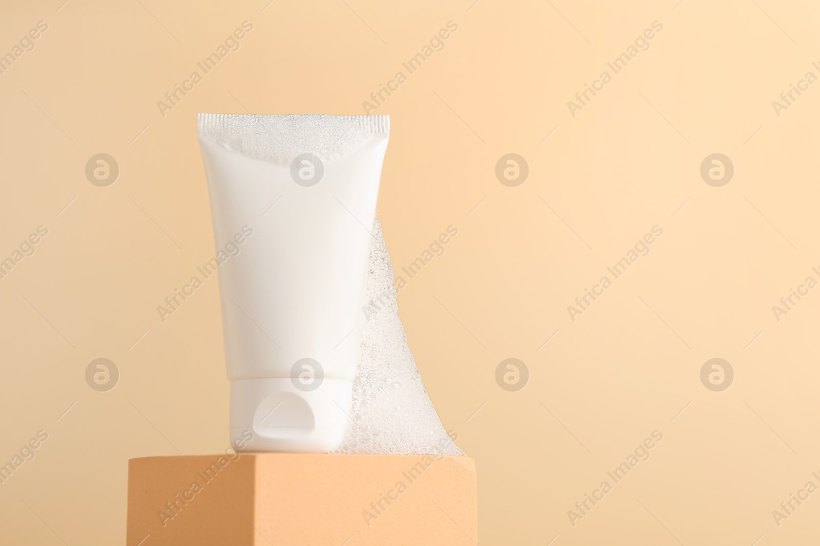 Photo of Tube of cosmetic product with foam on podium against beige background, space for text