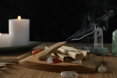 Photo of Smoldering palo santo stick, gemstones and burning candles on table, closeup