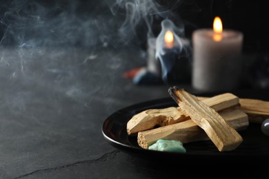 Photo of Smoldering palo santo stick, gemstone and burning candles on black table, closeup. Space for text