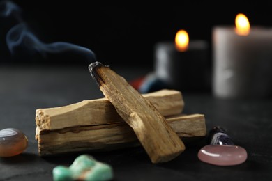 Photo of Smoldering palo santo stick, gemstones and burning candles on black table, closeup