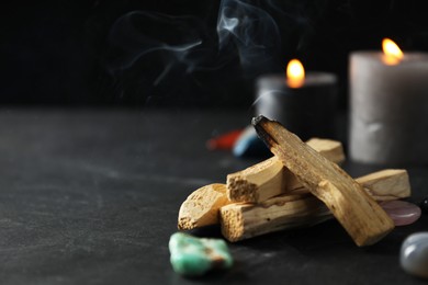 Photo of Smoldering palo santo stick, gemstones and burning candles on black table, closeup. Space for text