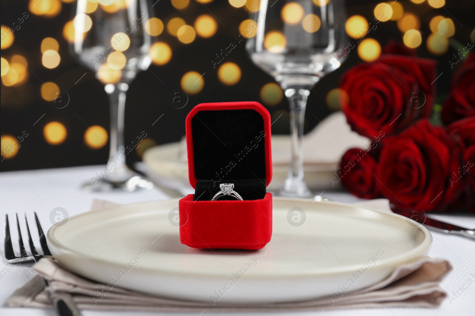 Photo of Engagement ring in box and tableware on white tablecloth, closeup. Table setting for romantic dinner