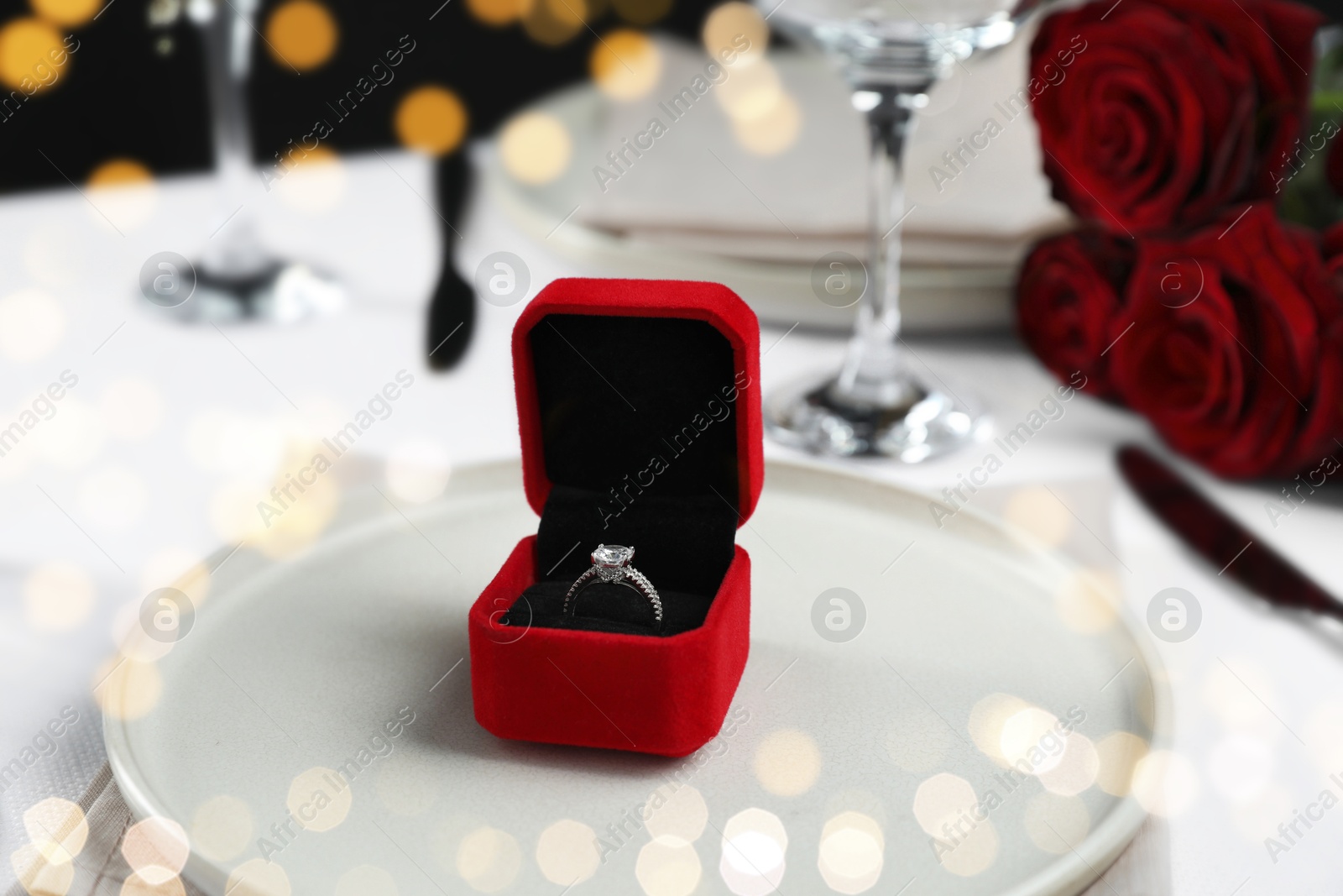 Photo of Engagement ring in box and plate on white tablecloth, closeup. Table setting for romantic dinner