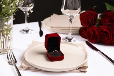 Engagement ring in box and tableware on white tablecloth, closeup. Table setting for romantic dinner