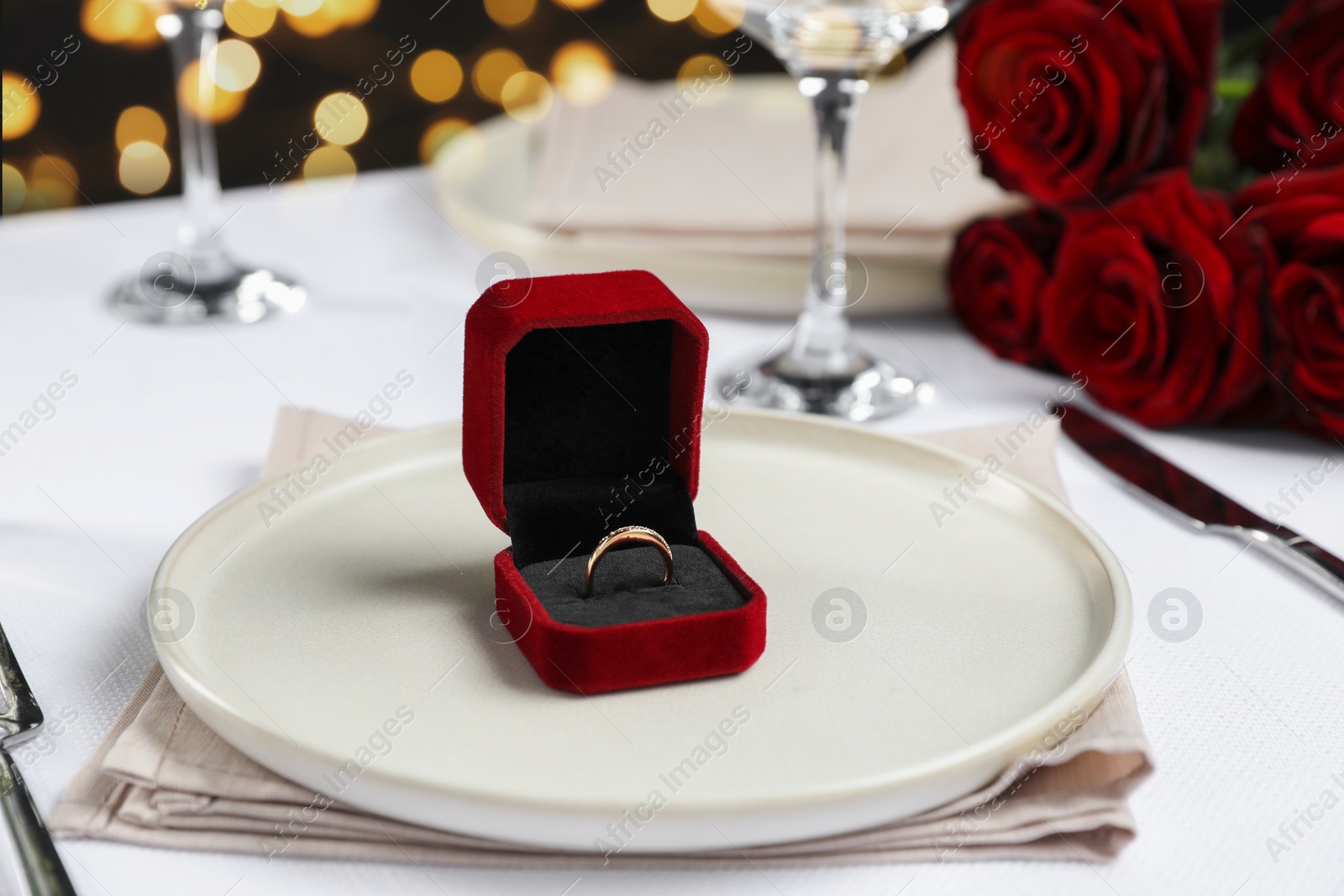 Photo of Engagement ring in box and tableware on white tablecloth, closeup. Table setting for romantic dinner