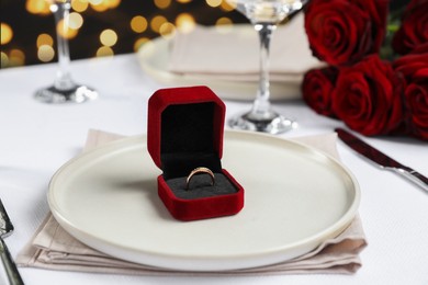 Engagement ring in box and tableware on white tablecloth, closeup. Table setting for romantic dinner