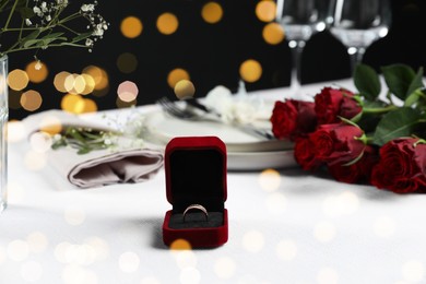 Photo of Engagement ring in box, flowers and tableware on white tablecloth. Romantic dinner