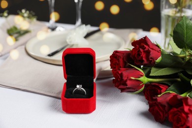 Engagement ring in box, rose flowers and tableware on white tablecloth, closeup. Romantic dinner