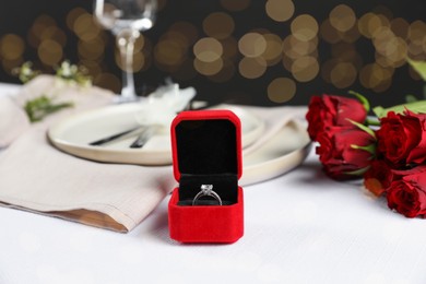 Engagement ring in box, rose flowers and tableware on white tablecloth, closeup. Romantic dinner