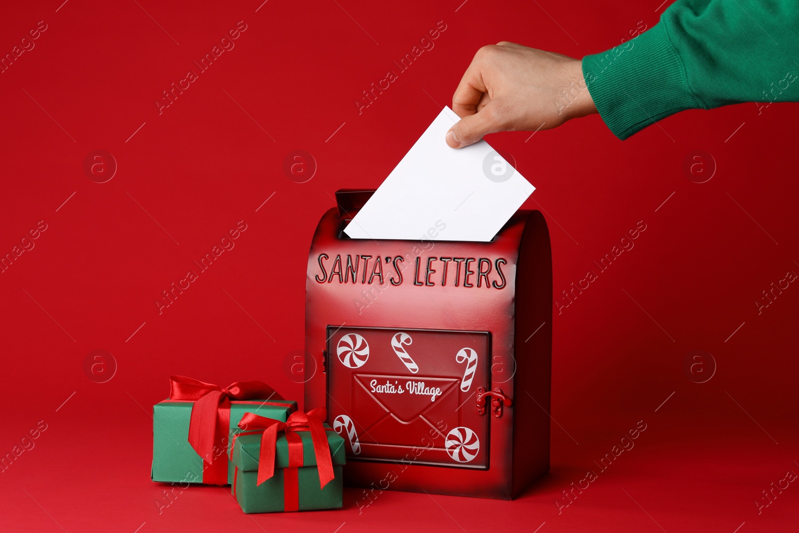 Photo of Man putting letter into Santa Claus mailbox against red background, closeup. Christmas tradition