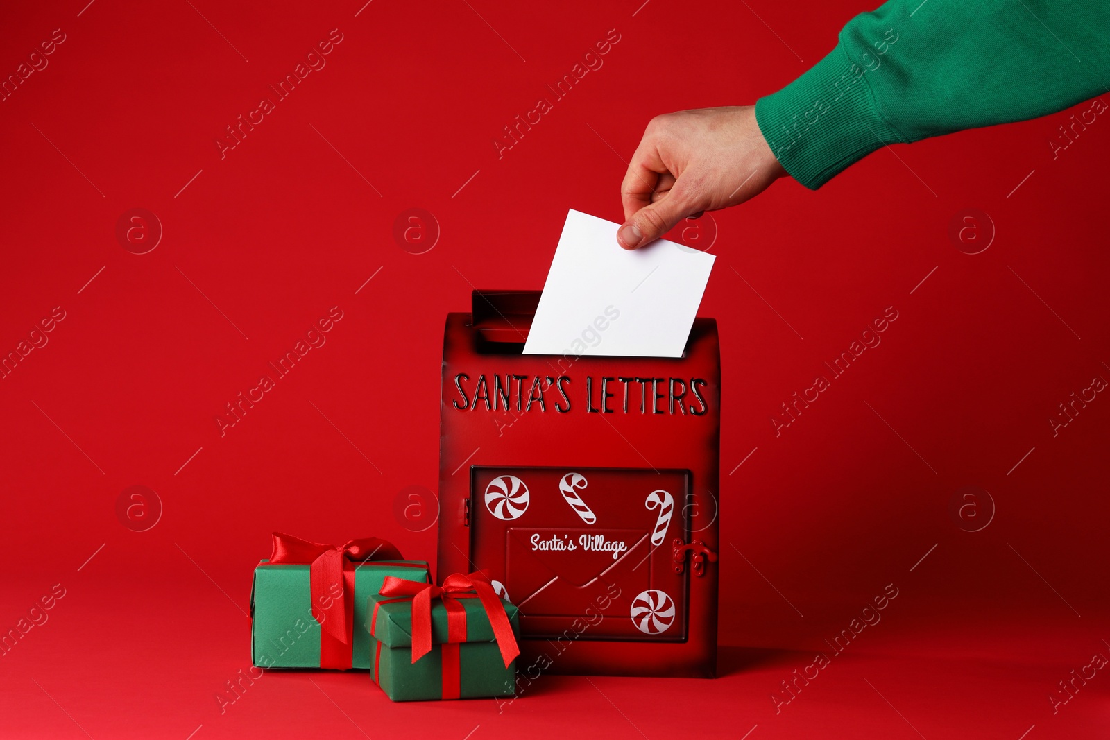 Photo of Man putting letter into Santa Claus mailbox against red background, closeup. Christmas tradition