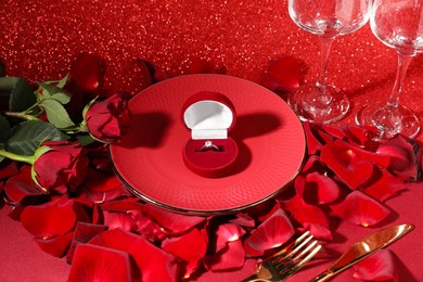 Engagement ring in box, tableware, rose flowers and petals on red glitter background. Table setting for romantic dinner