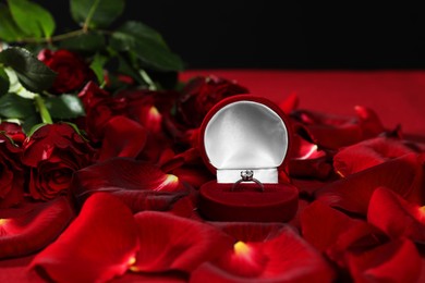 Engagement ring in box, rose flowers and petals on red table, closeup. Romantic proposal