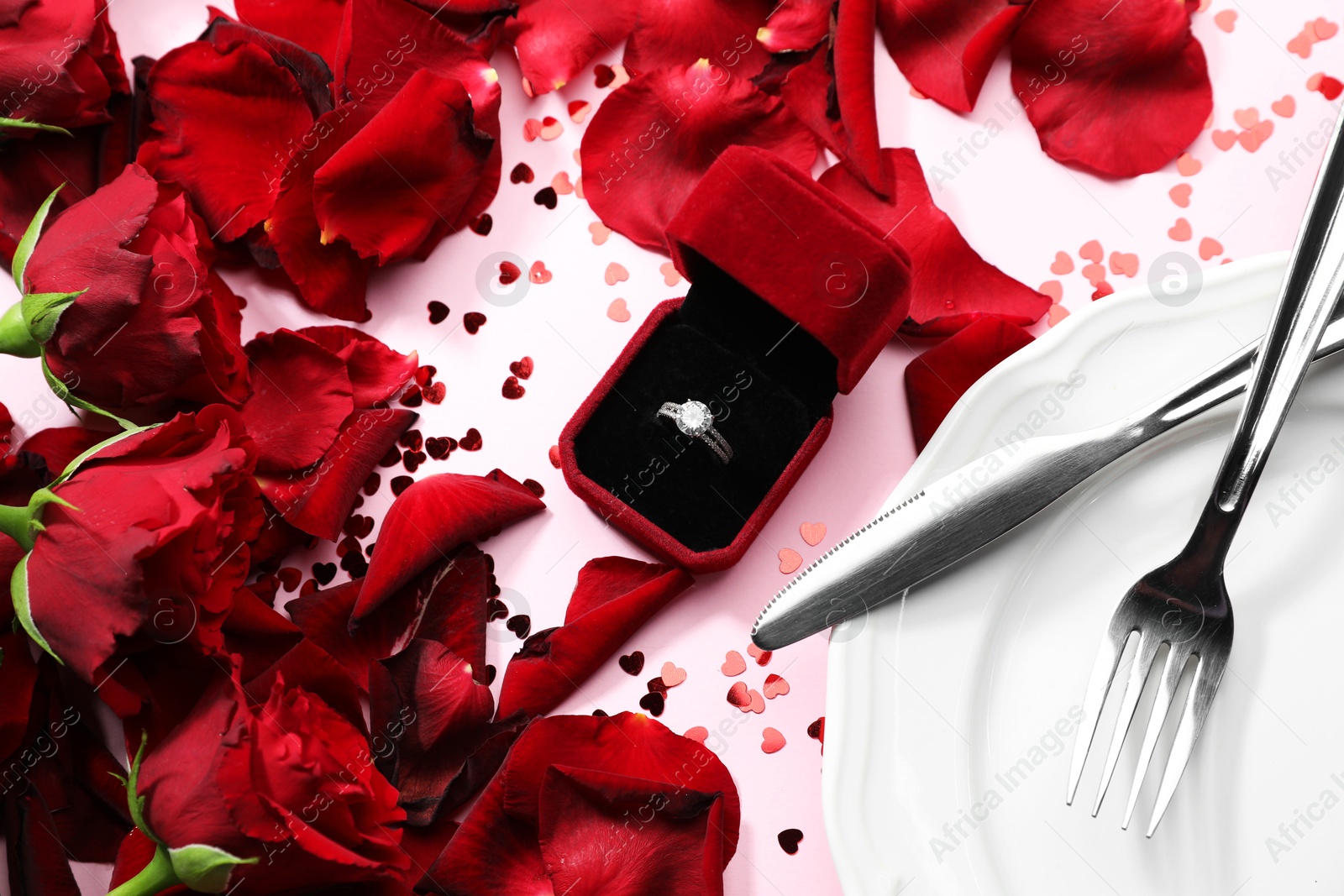 Photo of Beautiful setting for romantic dinner, engagement ring and rose petals on light pink table, flat lay