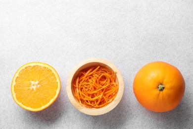 Photo of Fresh orange zest in bowl, whole and cut fruits on grey textured table, flat lay. Space for text