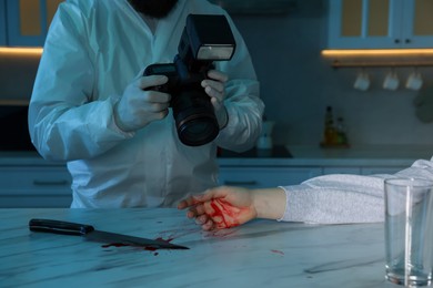 Photo of Forensic expert with camera, knife, blood and dead woman's body at countertop indoors, closeup