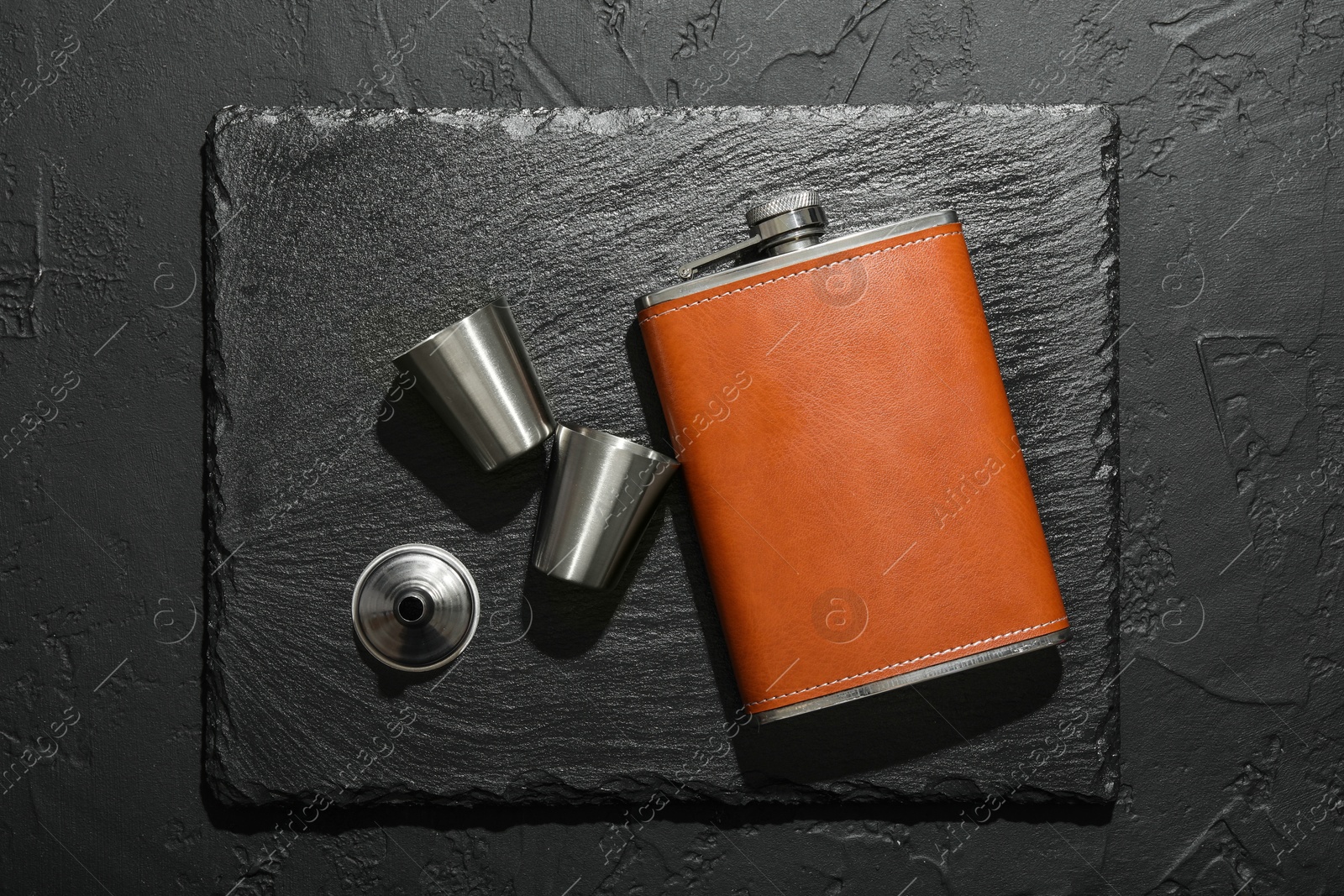 Photo of Hip flask, cups and funnel on black table, flat lay