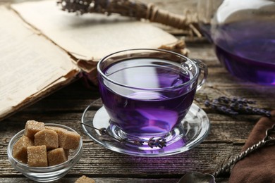 Photo of Aromatic lavender tea in glass cup, brown sugar, book and dry flowers on wooden table, closeup