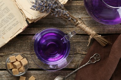 Photo of Aromatic lavender tea in glass cup, spoon, brown sugar, teapot, book and bunch of dry flowers on wooden table, flat lay