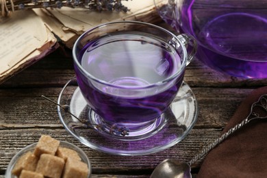 Photo of Aromatic lavender tea in glass cup, brown sugar, book, spoon and dry flowers on wooden table, closeup