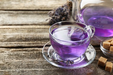 Photo of Aromatic lavender tea in glass cup, teapot, brown sugar and bunch of dry flowers on wooden table, closeup. Space for text