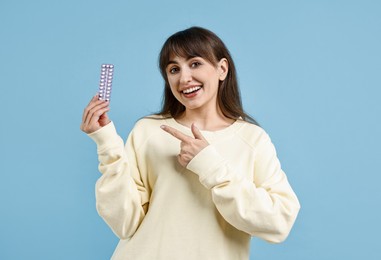 Smiling woman with blister of contraceptive pills on light blue background