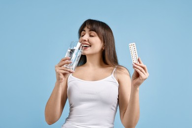 Young woman with contraceptive pills drinking water on light blue background