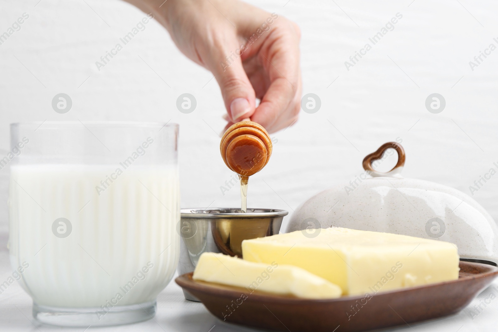 Photo of Woman with honey dipper, closeup. Fresh milk in glass and butter on white tiled table