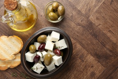 Photo of Marinated olives with feta cheese, bread pieces, oil and rosemary on wooden table, flat lay. Space for text