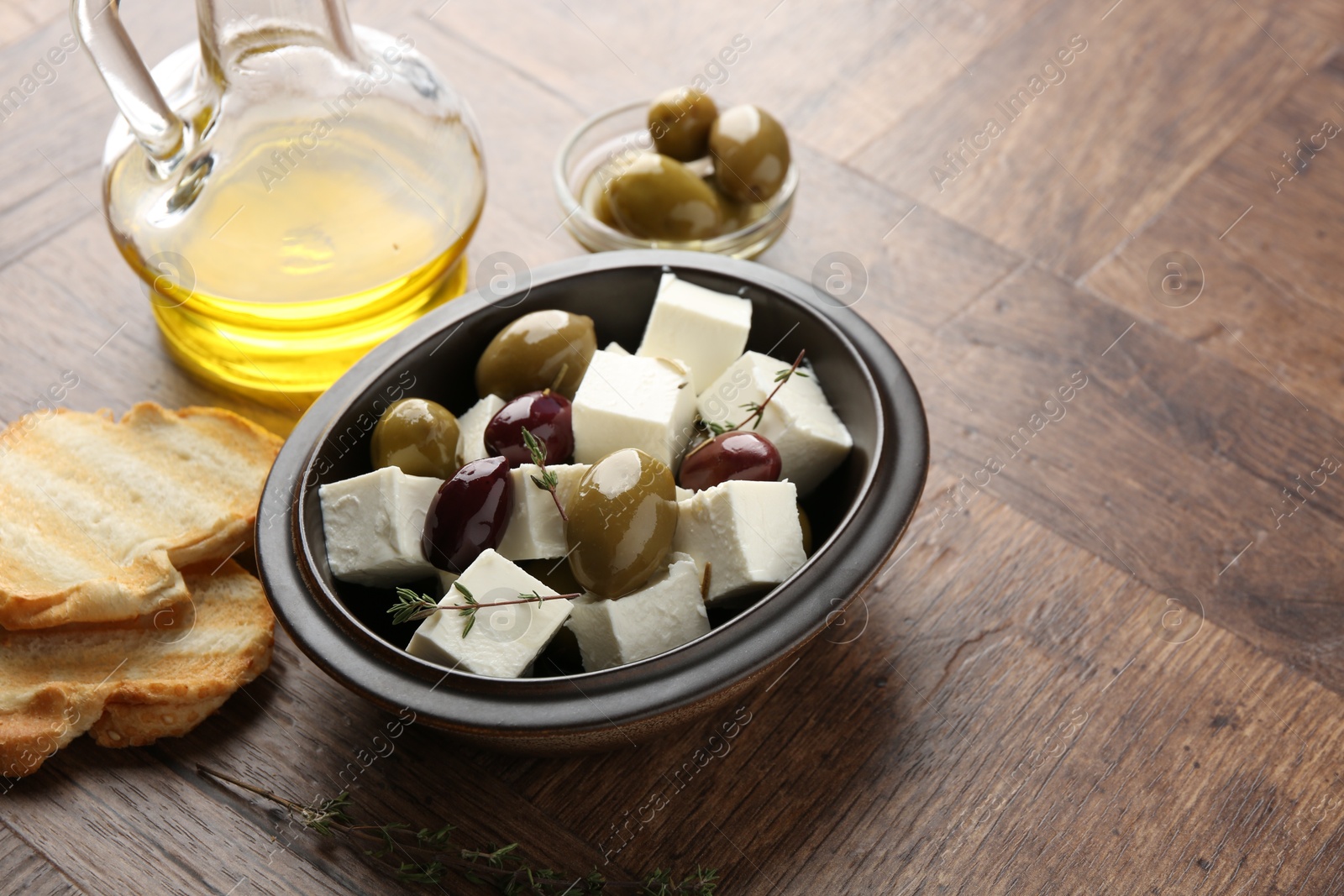 Photo of Marinated olives with feta cheese, bread pieces, oil and rosemary on wooden table, closeup. Space for text