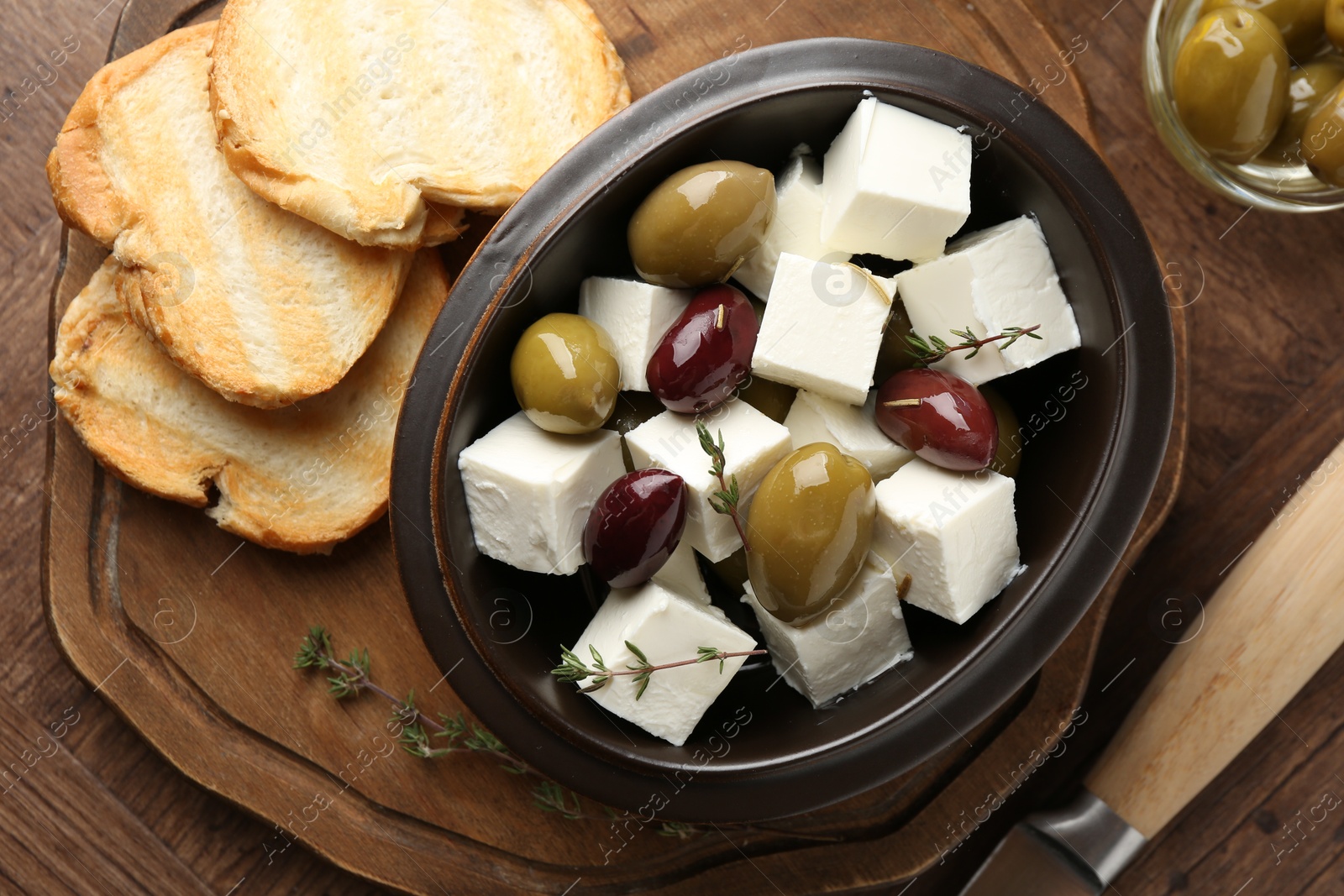 Photo of Marinated olives with feta cheese, bread pieces, oil and rosemary on wooden table, flat lay