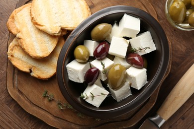 Photo of Marinated olives with feta cheese, bread pieces, oil and rosemary on wooden table, flat lay