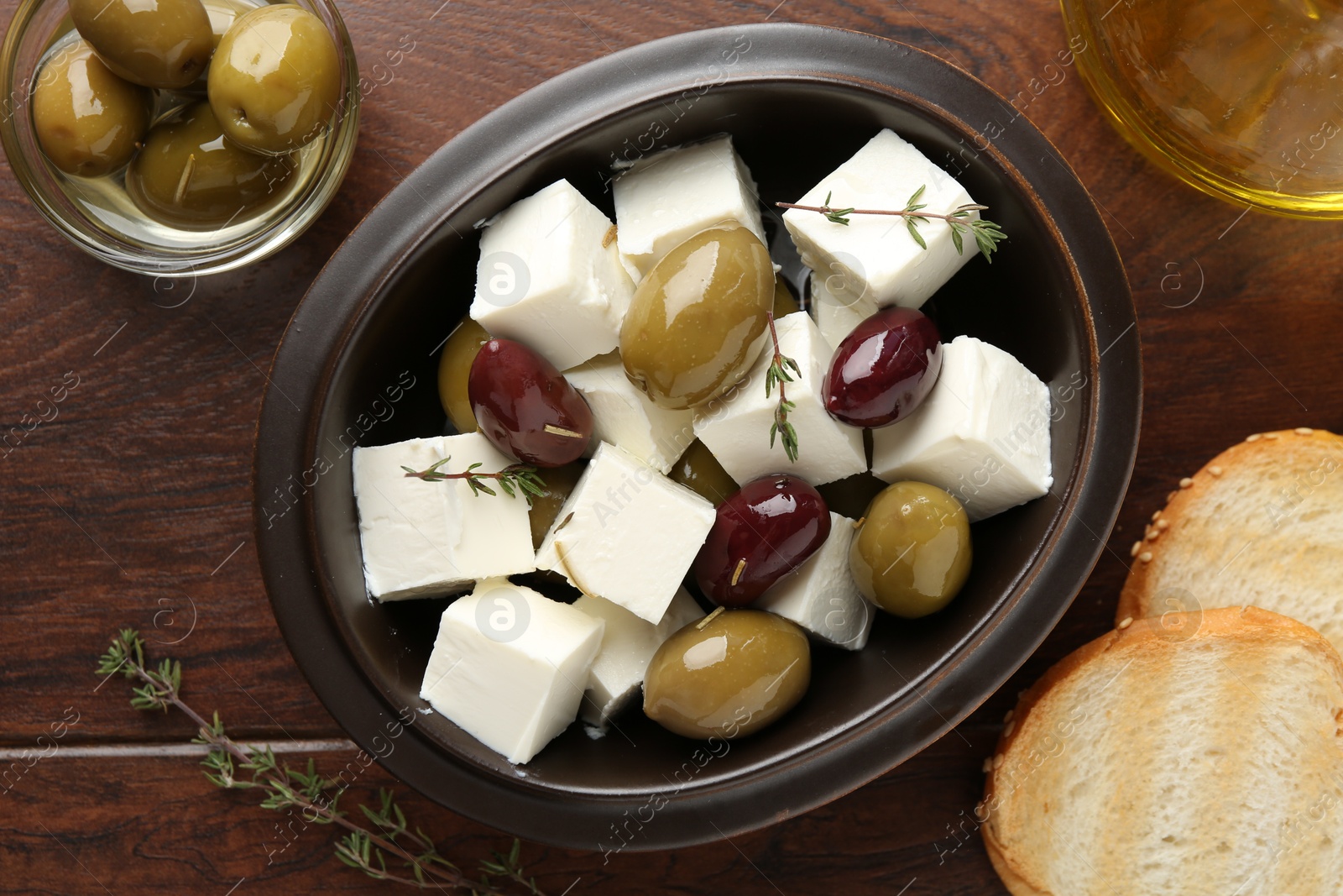 Photo of Marinated olives with feta cheese, bread pieces, oil and rosemary on wooden table, flat lay