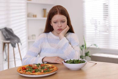 Sad teenage girl with pizza and bowl of salad at wooden table. Eating disorder