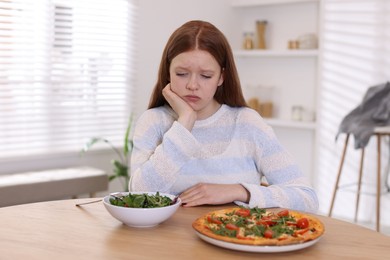 Photo of Sad teenage girl with pizza and bowl of salad at wooden table. Eating disorder