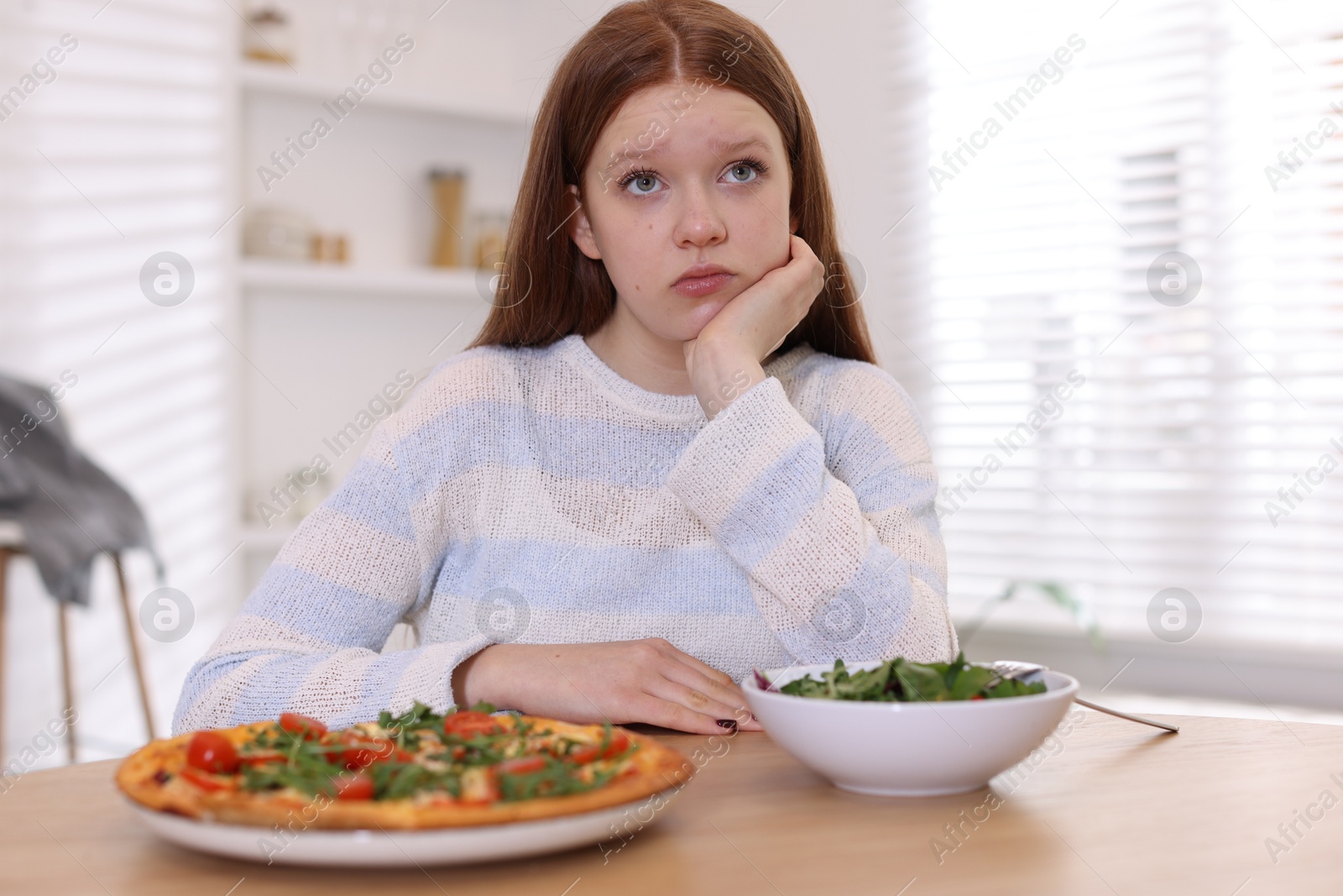 Photo of Sad teenage girl with pizza and bowl of salad at wooden table. Eating disorder