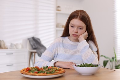 Sad teenage girl with pizza and bowl of salad at wooden table. Eating disorder