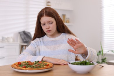 Photo of Sad teenage girl with pizza and bowl of salad at wooden table. Eating disorder