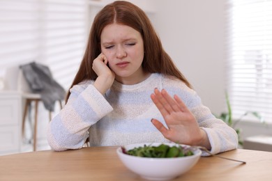 Photo of Sad teenage girl with bowl of salad at wooden table. Eating disorder