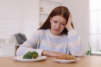 Sad teenage girl with broccoli and cookies at wooden table. Eating disorder