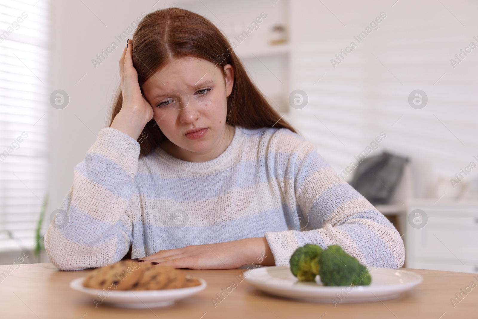 Photo of Sad teenage girl with broccoli and cookies at wooden table. Eating disorder