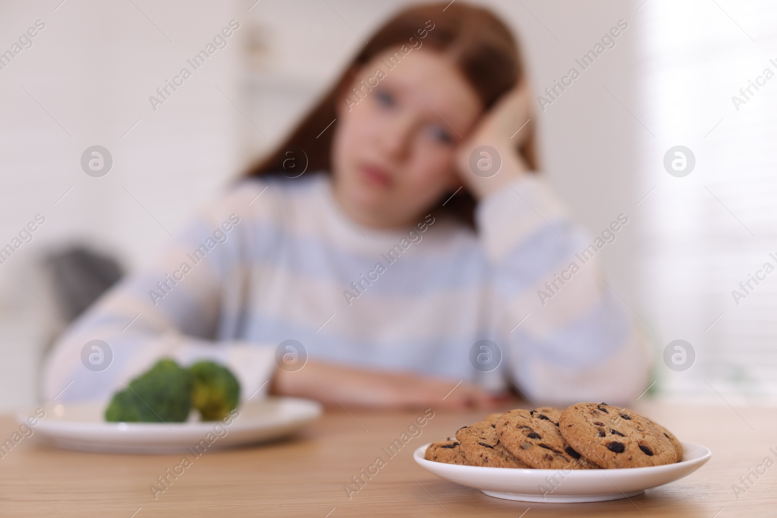 Photo of Sad teenage girl with broccoli and cookies at wooden table, selective focus. Eating disorder