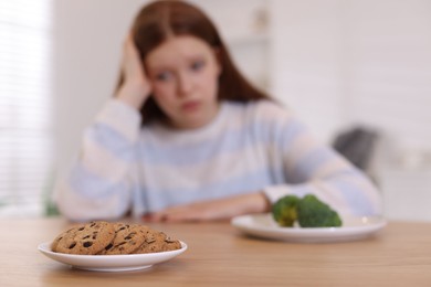 Photo of Sad teenage girl with broccoli and cookies at wooden table, selective focus. Eating disorder