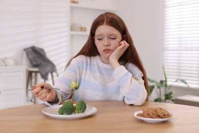 Sad teenage girl with broccoli and cookies at wooden table. Eating disorder