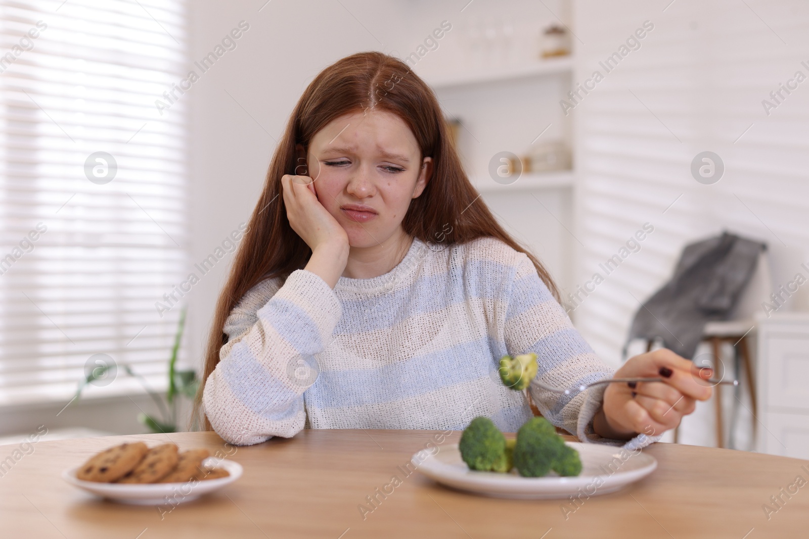 Photo of Sad teenage girl with broccoli and cookies at wooden table. Eating disorder