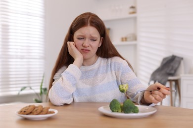 Photo of Sad teenage girl with broccoli and cookies at wooden table. Eating disorder