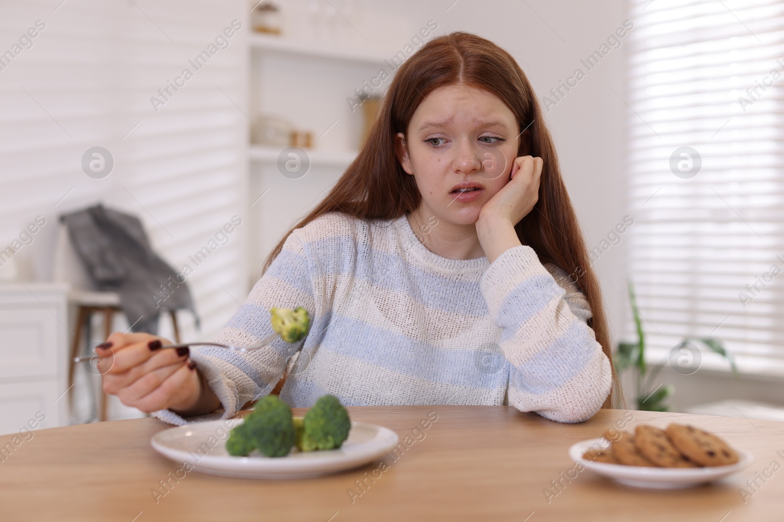 Photo of Sad teenage girl with broccoli and cookies at wooden table. Eating disorder