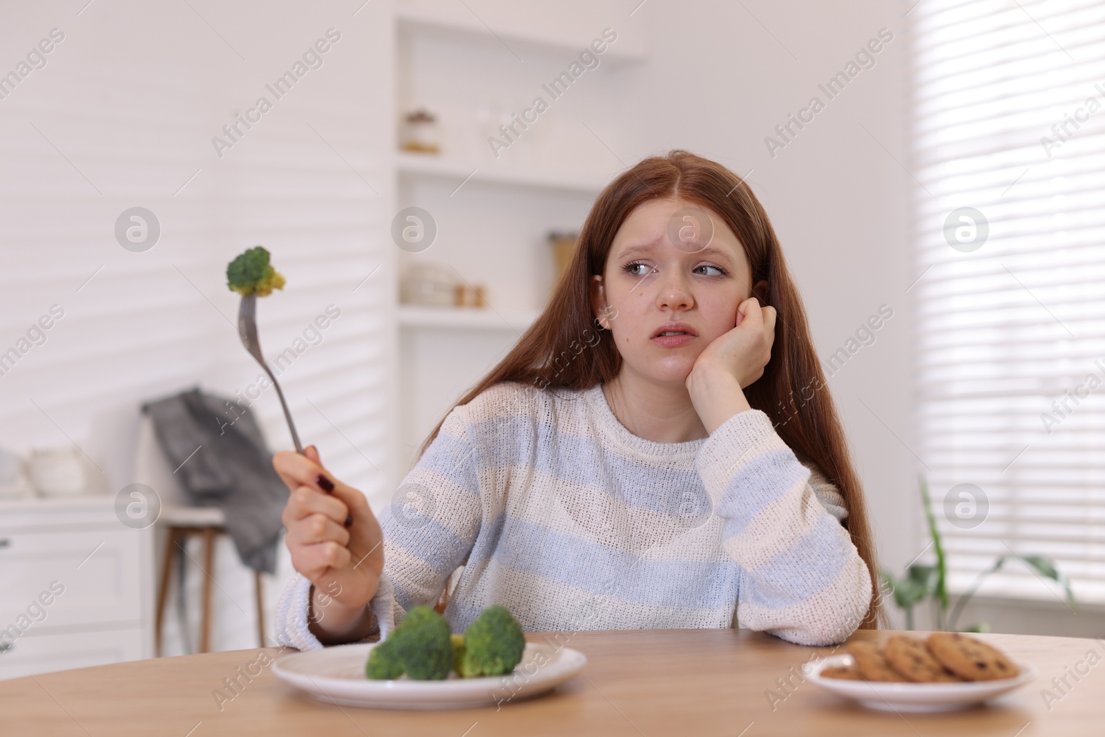 Photo of Sad teenage girl with broccoli and cookies at wooden table. Eating disorder