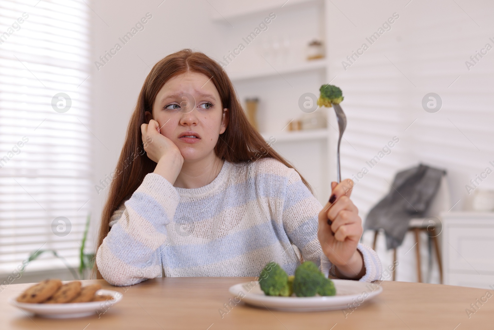 Photo of Sad teenage girl with broccoli and cookies at wooden table. Eating disorder