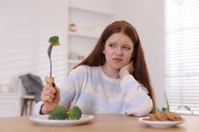 Photo of Sad teenage girl with broccoli and cookies at wooden table. Eating disorder